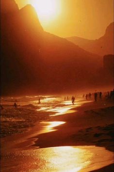 people are walking on the beach as the sun goes down in the mountains behind them