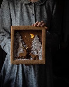 a person holding up a small wooden frame with deer and pine trees on it in the dark