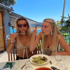 two women sitting at a table with food in front of their faces and one is eating