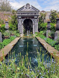 an outdoor garden with a pond surrounded by greenery and stone arches, along with blue flowers