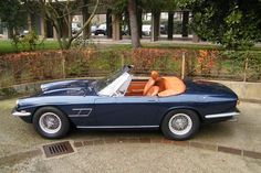 an old blue sports car parked in front of a building with trees and bushes behind it