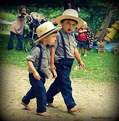 two young boys wearing hats and suspenders walking down a dirt road with people in the background