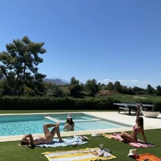 two women lounging in front of a swimming pool with towels on the grass near it