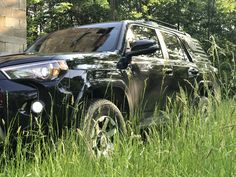 a black suv parked in tall grass next to a brick wall and trees on the other side