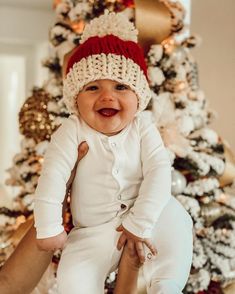 a smiling baby wearing a red and white knitted hat sitting in front of a christmas tree