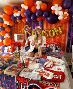 a woman sitting on top of a bed surrounded by orange and purple balloons in the background