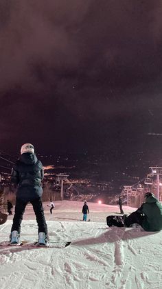 two people standing on top of a snow covered slope at night with ski lift in the background