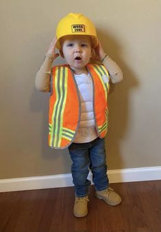 a little boy wearing a construction vest and hat with his hands on his head while standing in front of a wall