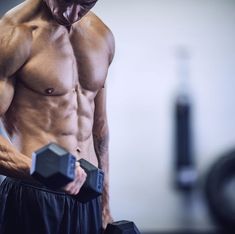 a shirtless man holding two dumbbells in his right hand and looking at the camera