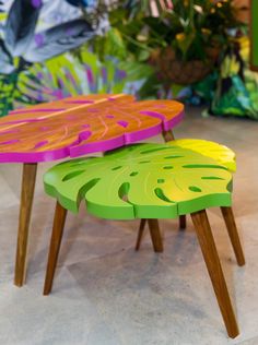 two colorful tables sitting next to each other on top of a cement floor with plants in the background