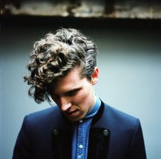 a man with curly hair wearing a blue shirt and black blazer looking down at his cell phone