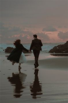 a man and woman holding hands while walking on the beach at sunset with their reflection in the wet sand