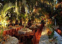 an outdoor dining area with tables and chairs covered in orange linens, surrounded by greenery