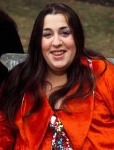 a woman in an orange jacket sitting on a bench smiling at the camera with her hands behind her back