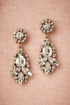 two pairs of earrings are shown on a pink surface with white and gold accents, one is