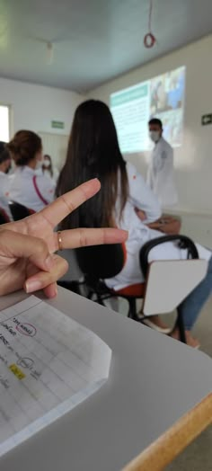 a person making a peace sign in front of other people sitting at desks and chairs