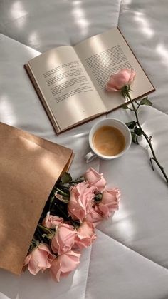 an open book and some pink flowers on a white table cloth with a cup of coffee