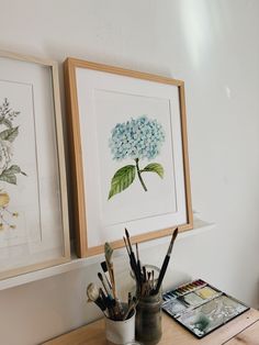 two framed pictures on the wall above a desk with paintbrushes and watercolors