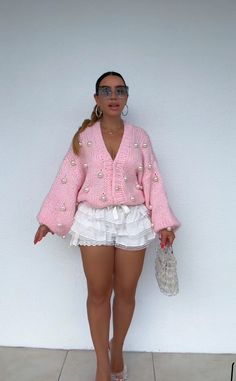 a woman standing in front of a white wall wearing a pink sweater and white skirt