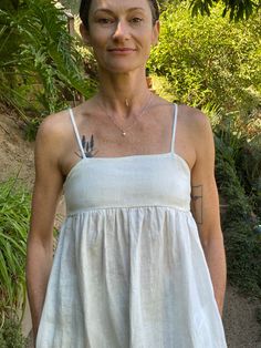 a woman wearing a white dress standing in front of some bushes and trees with a tattoo on her arm