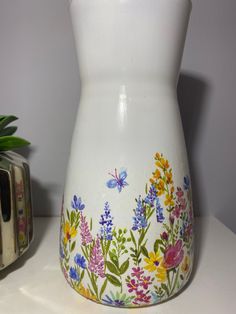 a white vase with colorful flowers painted on it next to a small potted plant