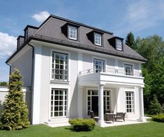 a large white house with black roof and windows on the top floor, sitting in front of a lush green lawn