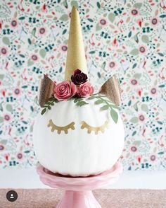 a white pumpkin decorated with flowers and a unicorn's horn on top, sitting in front of a floral wallpaper