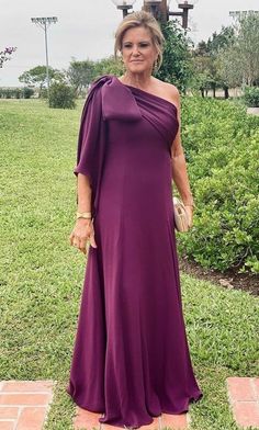 an older woman in a purple dress standing on the side of a brick walkway with grass and bushes behind her