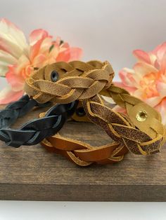 three braided leather bracelets sitting on top of a wooden board with flowers in the background