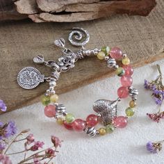 This artisan handmade double-strand bracelet showcases the natural beauty of beautiful grade gemstones, including Strawberry Quartz (10.8mm), Rutilated Quartz (6.8 mm), and Prehnite (8 mm), that carefully selected to make the colors of this bracelet stand out, creating a harmonious blend of pink, green, and yellow hues. The bracelet is adorned with intricate Karen Hill Tribe fine silver accents, featuring charming elements like a hammered heart-shaped bead, flower S-clasp, and unique charms, Including 925 sterling silver, all expertly strung together with a strong and durable waxed cord, finished with woven ends and Hill Tribe fine silver chain. Whether you're looking to add a statement piece to your jewelry collection, attend a festival or a boho-themed event, search for a thoughtful gift Bohemian Beaded Tourmaline Jewelry, Bohemian Tourmaline Jewelry With Natural Stones, Bohemian Tourmaline Bead Jewelry, Bohemian Tourmaline Round Beads Jewelry, Handmade Tourmaline Bracelet, Tourmaline Gemstone Beads Bracelets For Jewelry Making, Spiritual Tourmaline Beaded Bracelets With Natural Stones, Double Strand Gemstone Beads Bracelet As Gift, Spiritual Double Strand Gemstone Bracelets