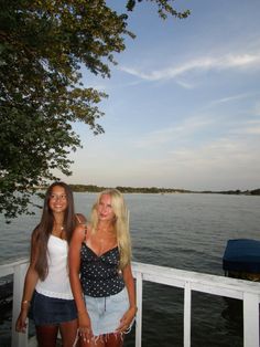 two beautiful young women standing next to each other on a pier near the water and trees