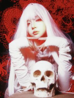 a woman with long white hair sitting in front of a skull and holding her hand up to her face