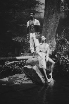 an old photo of two people sitting on some rocks in the water near a tree