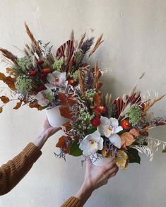 two hands holding flowers in front of a white wall with leaves and grass on it