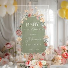 a baby shower sign sitting on top of a wooden stand with flowers and balloons in the background