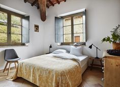 a bed sitting under two windows in a bedroom next to a wooden dresser and chair