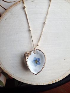 Capture the fleeting beauty of these lovely blooms forever in these unique pressed flower pendant necklaces. Genuine pressed Forget-Me-Nots are set onto a genuine little clam shell collected along the beaches near Tampa Bay. The inside of each clam is painted white and then the flower is set. We apply numerous coats of resin to seal the bloom safely inside and then we gild the edges and the back in a gold paint. No little clams were left homeless by our collection..these are all naturally occurr Handmade Flower Shell Necklace Gift, White Birth Flower Keepsake Jewelry, White Birth Flower Jewelry Keepsake, White Birth Flower Jewelry For Keepsake, Nature-inspired White Birth Flower Necklace, Nature-inspired White Necklace With Pressed Flowers, White Nature-inspired Necklace With Pressed Flowers, Nature-inspired White Jewelry With Birth Flower, White Nature-inspired Jewelry With Birth Flower