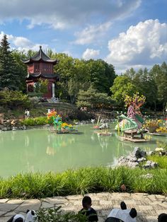 people are sitting on the ground looking at a pond with animals and flowers in it