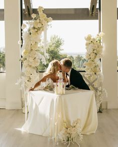 a bride and groom kissing in front of a table with white flowers on it at their wedding reception