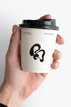 a hand holding a coffee cup with a black lid and logo on the bottom, in front of a white background