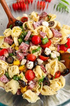 a pasta salad in a glass bowl with a wooden spoon and tomatoes on the side