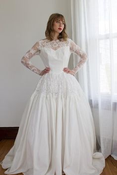 a woman in a white wedding dress standing by a window with her hands on her hips