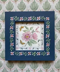 a blue frame with pink flowers and green leaves on the wall next to a flowered wallpaper