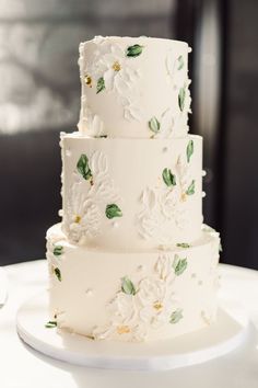 a three tiered white wedding cake with green leaves and flowers on the bottom layer