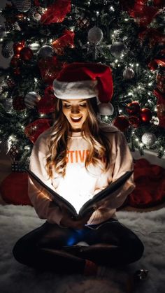 a woman is sitting in front of a christmas tree and reading a book while wearing a santa hat