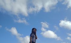 a woman standing on top of a lush green field under a blue sky with clouds