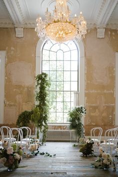 a room with chairs, tables and flowers in front of a large window that has a chandelier hanging from the ceiling