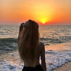 a woman standing on top of a sandy beach next to the ocean at sunset with her hair blowing in the wind