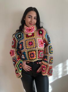 a woman standing in front of a wall wearing black pants and a colorful crocheted sweater