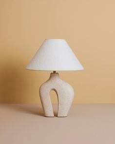 a white ceramic table lamp with a light shade on it's base and a beige wall in the background